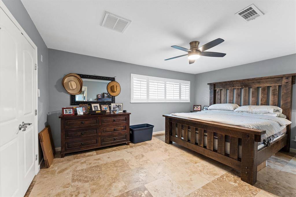 This bedroom features a spacious layout with a large wooden bed, stylish tile flooring, and a ceiling fan. It has a dresser with a mirror, ample natural light from a window with shutters, and a neutral color scheme for a cozy atmosphere.