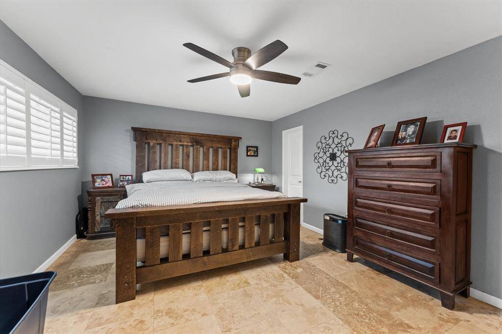 This bedroom features a cozy, well-lit space with neutral walls and a ceiling fan. It includes a large bed with a decorative comforter, a window with shutters offering natural light, and a white bedside table. The room is finished with tile flooring, providing a clean and modern look.