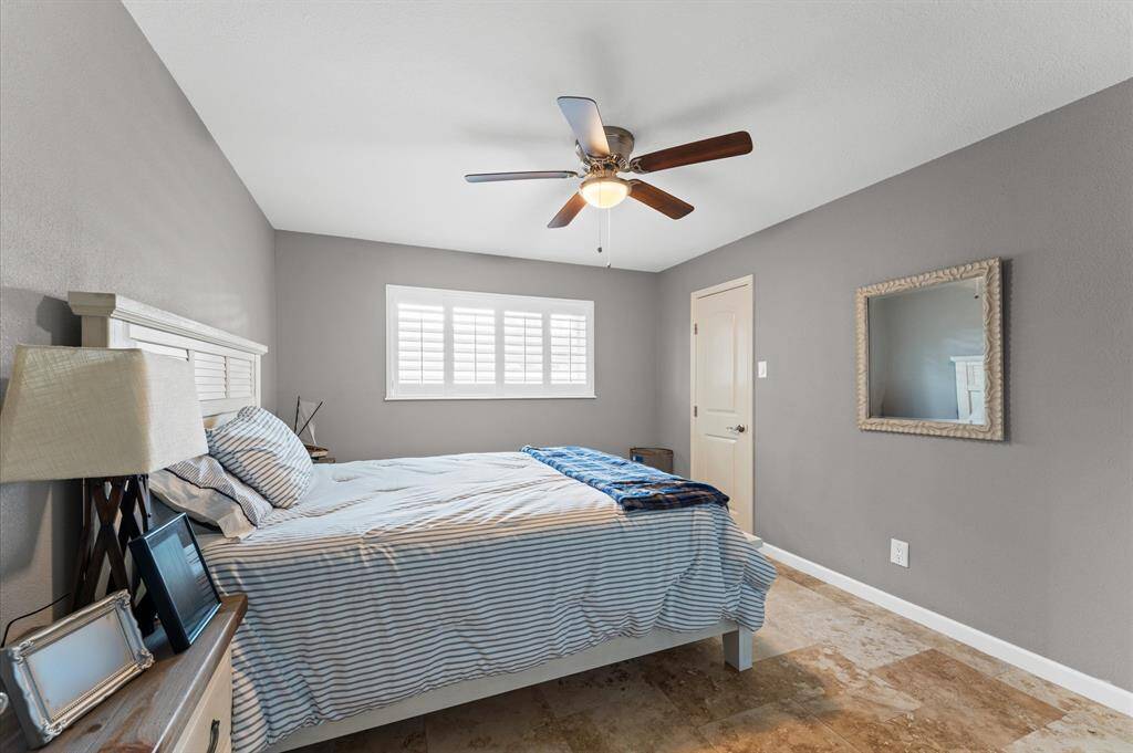 This great size bedroom features neutral gray walls, a ceiling fan, and ample natural light from a window with shutters. The room includes a comfortable bed, a bedside table, and a wall mirror, creating a welcoming atmosphere.