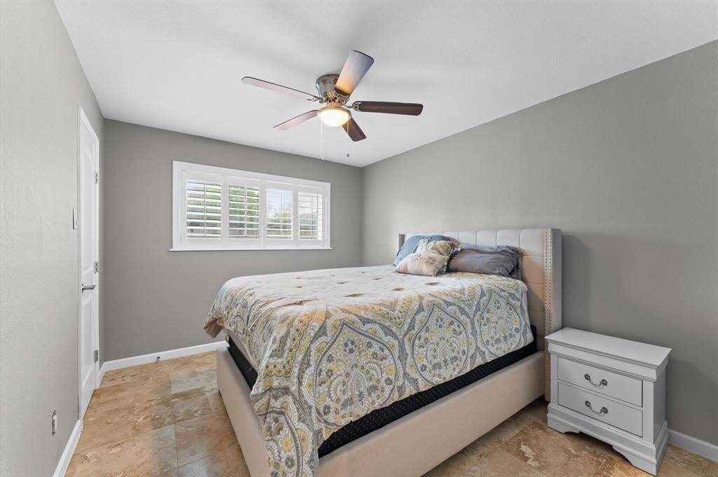 This cozy bedroom features a ceiling fan, neutral gray walls, and a large window with shutters for natural light. It includes a bed, a wooden table, and ample space for personal items or hobbies.