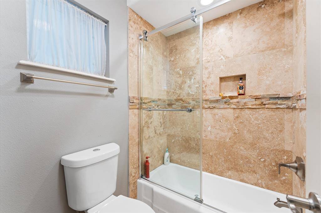 This bathroom features a modern design with a glass-enclosed bathtub and shower, elegant stone tile walls, and a small built-in shelf. It includes a window for natural light and a sleek, neutral color palette.