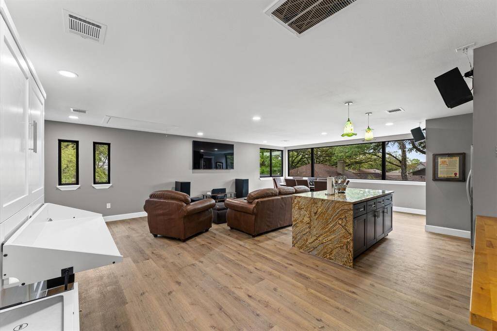 The spacious apartment features a modern open-plan design with large windows offering ample natural light. It includes a cozy seating area with leather sofas, a dining table, a Murphy bed and a stylish kitchen with a stone backsplash and dark wood cabinets, perfect for entertaining. The wood flooring adds warmth and elegance to the space.