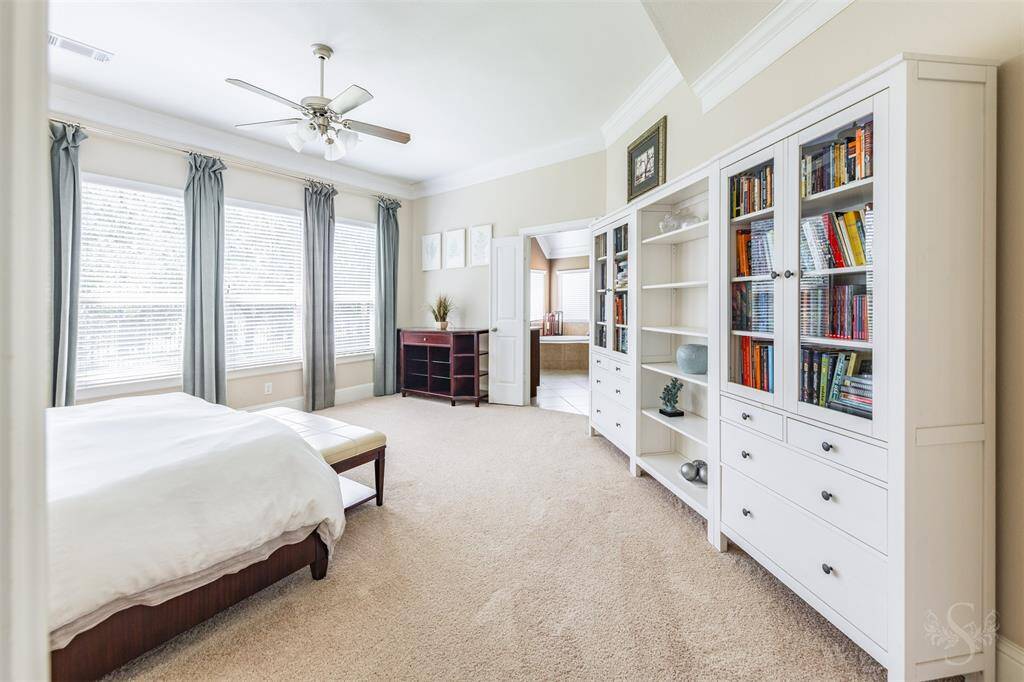 The spacious primary suite features plush carpet, a high ceiling with crown molding, and a wall of windows filling the room with natural light. A peaceful retreat with plenty of space for your king size bed and a sitting area by the windows.