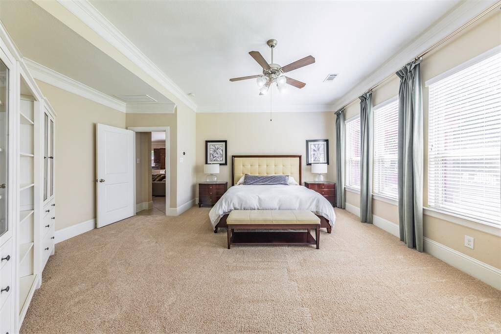 The spacious primary suite features plush carpet, a high ceiling with crown molding, and a wall of windows filling the room with natural light. A peaceful retreat with plenty of space for your king size bed and a sitting area by the windows.