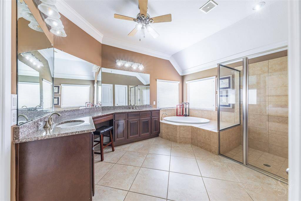 The spa-inspired primary bath includes a deep soaking tub, enclosed shower with tile surround, and an extended granite vanity with double sinks. Elevated ceilings with crown molding, recessed lighting and elegant tile floors complete the space.