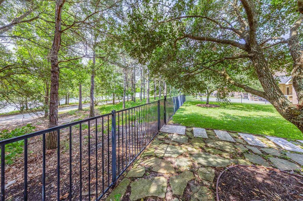 A scenic walking path and lake view lie just beyond the wrought iron fence, offering a serene backdrop framed by shady trees. The stone pathway and open yard create a charming transition from backyard to nature.