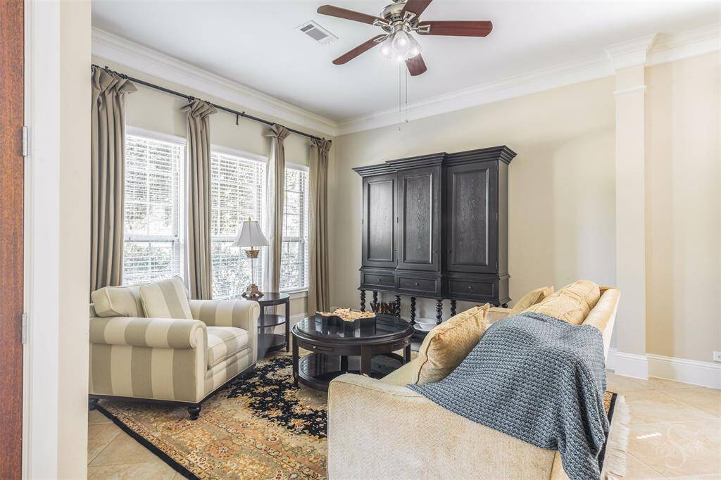A cozy sitting area in the living room is bathed in sunlight from the surrounding windows, offering a peaceful spot to relax. Neutral tile flooring and crown molding add to the room’s refined atmosphere.