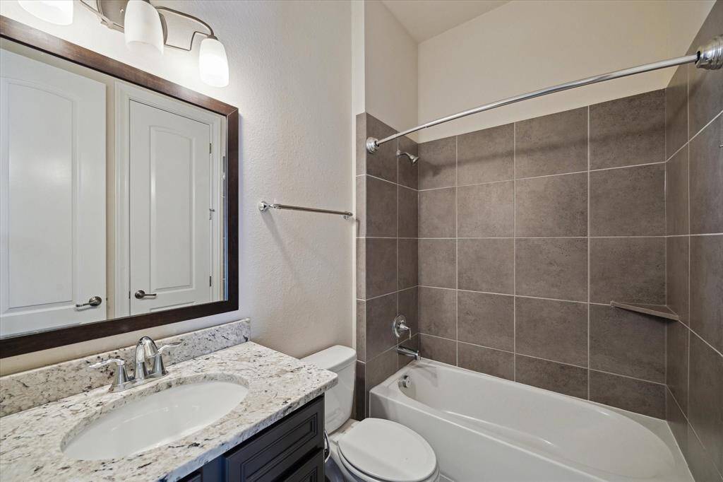 This bathroom features modern finishes with a granite countertop, sleek dark vanity, and large mirror. The bathtub is surrounded by stylish gray tile, and overhead lighting adds brightness.