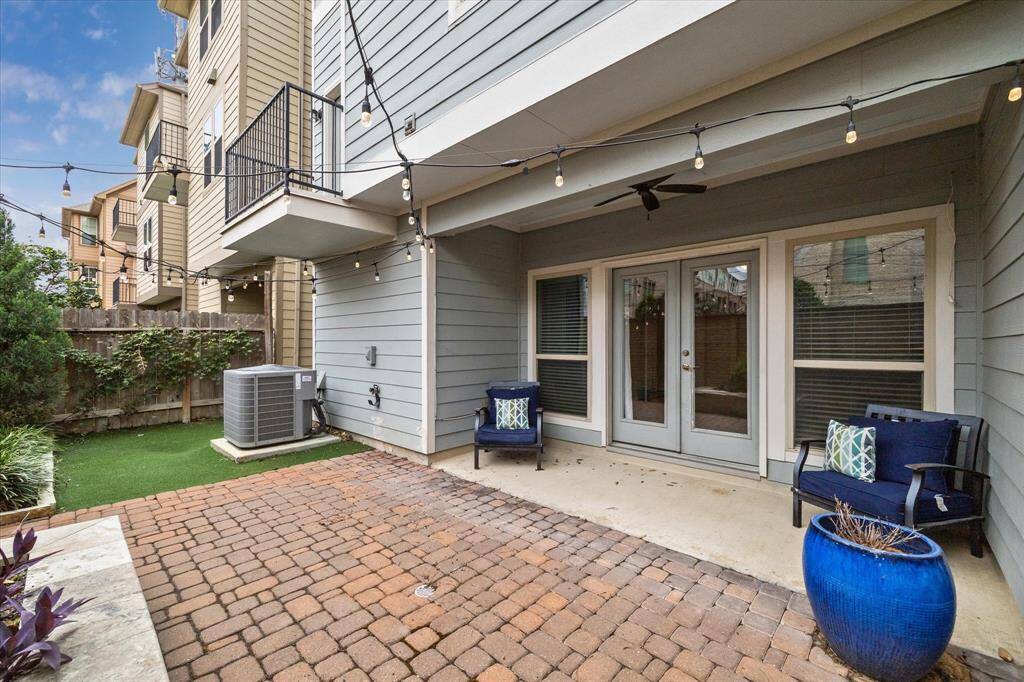 Covered backyard patio with string lights, a paver flooring, and a small seating area. The area includes artificial grass and a fence for privacy, making it perfect for relaxing or entertaining.