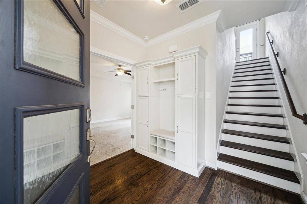 This inviting entryway features elegant hardwood floors, a built-in storage unit with cabinets and shoe cubbies, and a staircase leading upstairs. A glimpse of a bright, open living area with a ceiling fan is visible, creating a welcoming first impression.