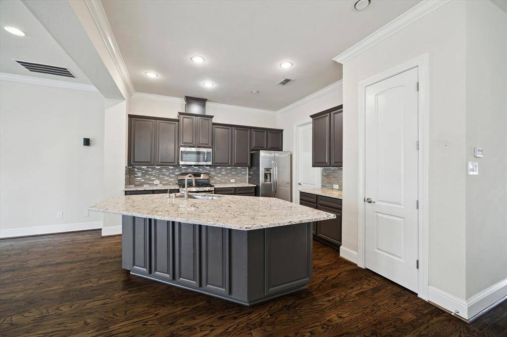 This kitchen features dark cabinetry with a modern backsplash, granite countertops, and a central island. Stainless steel appliances and hardwood floors complete the sleek, contemporary look.