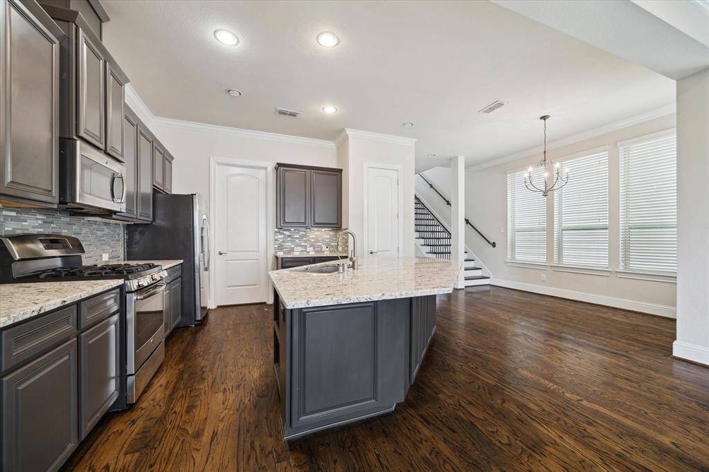 Modern kitchen with dark cabinetry, stainless steel appliances, and a granite countertop island. The open layout flows into a dining area with hardwood floors and large windows, providing ample natural light. A staircase leads to the upper level, adding to the spacious feel.