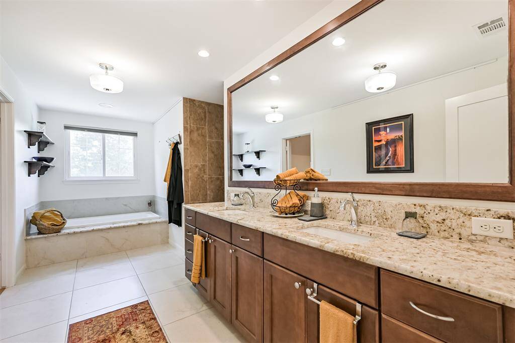 This stunning primary bathroom offers a peaceful, spa-like feel with double sinks, granite countertops, warm wood-framed mirror, and abundant storage. Recessed lighting and natural light make the space feel bright and welcoming. Relax and unwind in the oversized soaking tub or step into the luxurious walk-in shower, complete with floor-to-ceiling tile. Thoughtful details and warm accents throughout create a perfect balance of beauty and comfort.