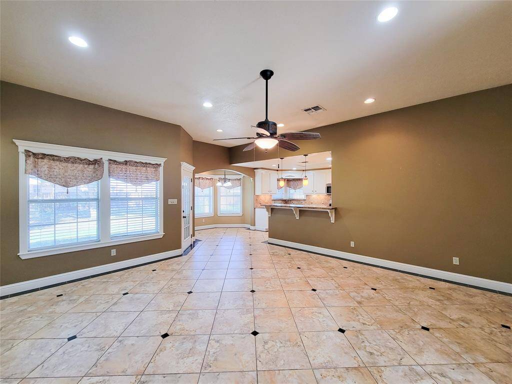 This spacious, open-concept living area features tiled floors, ample natural light from large windows, and a ceiling fan. It seamlessly connects to the kitchen, which includes a breakfast bar and pendant lighting, making it ideal for entertaining.