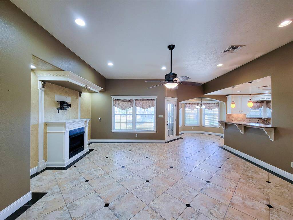 This spacious living area features tiled floors, a cozy fireplace, and recessed lighting. It includes a ceiling fan and a breakfast bar that opens to the kitchen. Large windows and a door to the outside provide ample natural light.