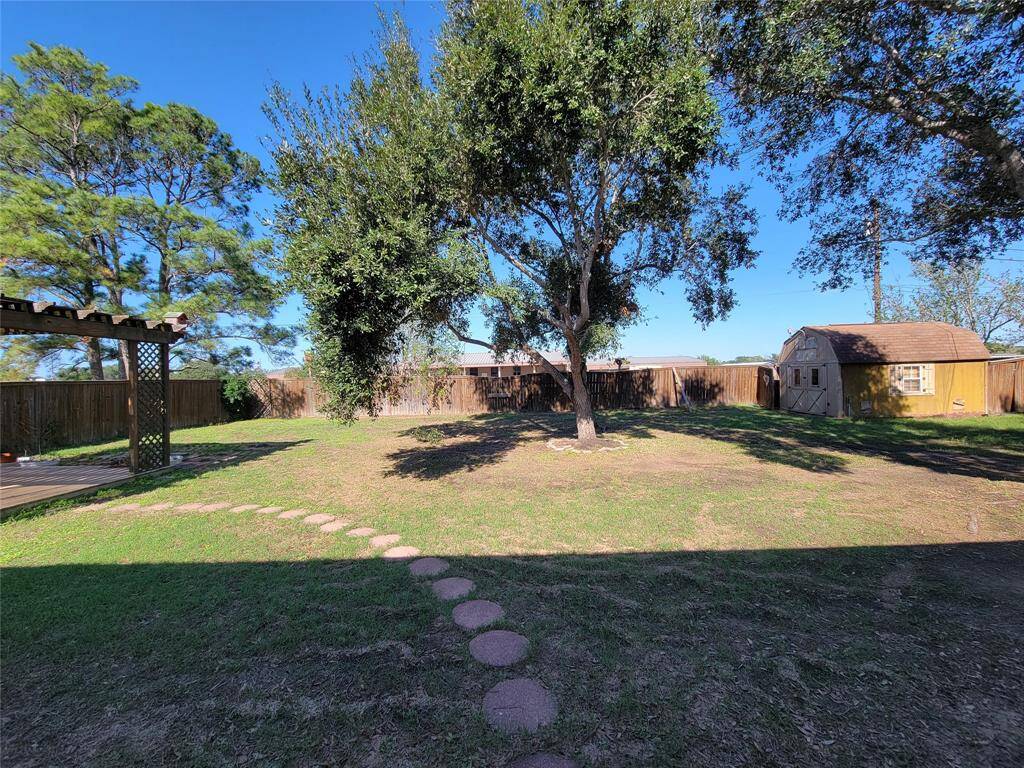 This backyard features a spacious, fenced area with mature trees and a small shed. A pergola-covered patio and a stepping stone path add charm and functionality, ideal for outdoor activities and relaxation.