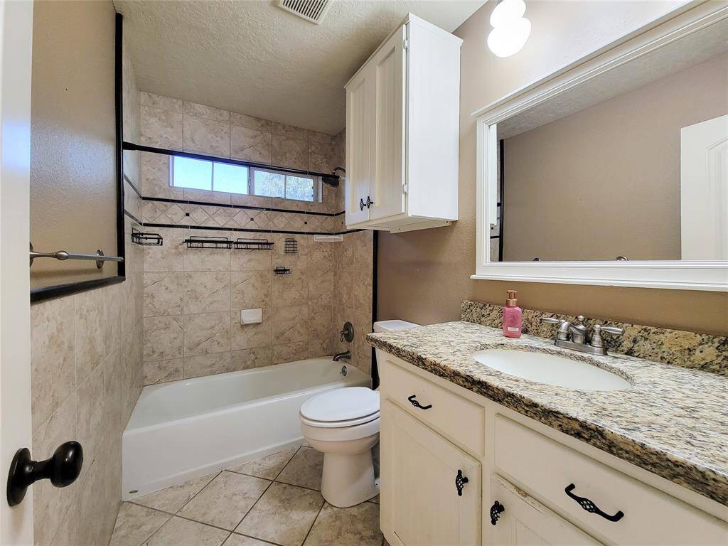 This bathroom features a granite countertop with a sink, white cabinetry, and a tiled bathtub/shower combo. The neutral tile flooring and wall tiles add a classic touch, while a large mirror and overhead lighting enhance the space.