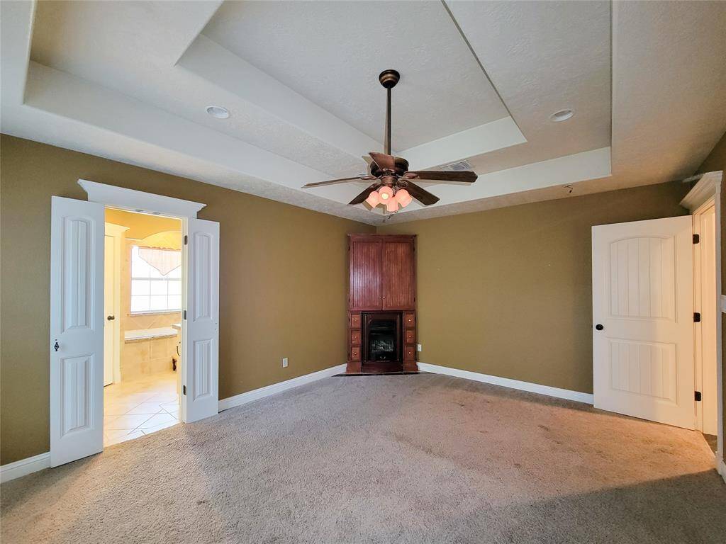 This spacious room features a tray ceiling with a ceiling fan, carpeted floors, and warm wall colors. It includes double doors leading to a well-lit bathroom with a large window, and a cozy built-in fireplace and cabinet.