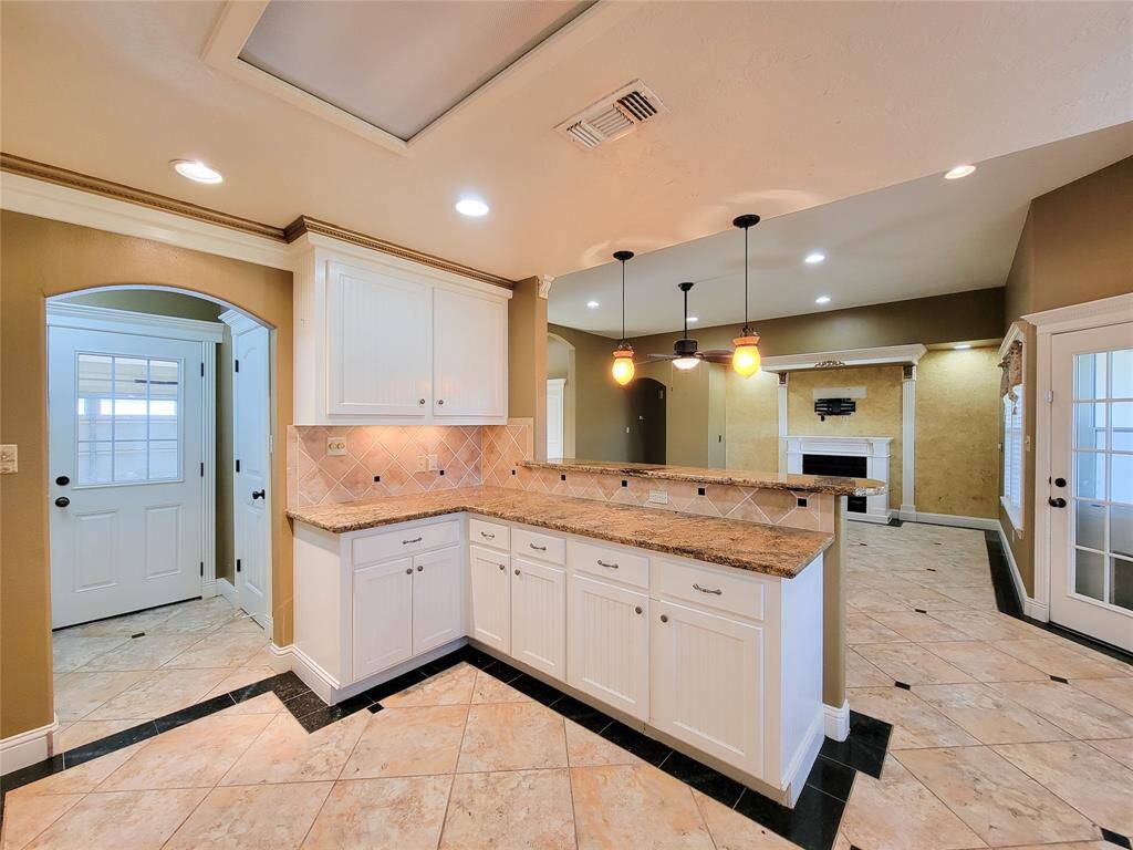 This is a spacious, open-concept kitchen with white cabinetry, granite countertops, and modern pendant lighting. It features tile flooring and has a view of the adjoining living area with a fireplace. There are multiple doors leading to other rooms, including one with glass panels allowing for natural light.