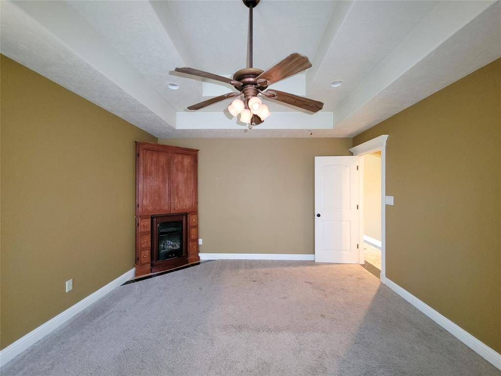 This is a spacious room with neutral carpeting, warm-toned walls, and a wooden entertainment unit with a built-in fireplace. The room features a ceiling fan with lighting and has a door leading to another area of the house.