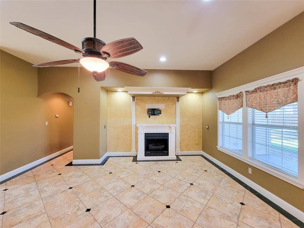 This photo shows a spacious room with tiled flooring and warm neutral colors. It features a ceiling fan with lighting, a cozy fireplace, and large windows with curtains, allowing for plenty of natural light.
