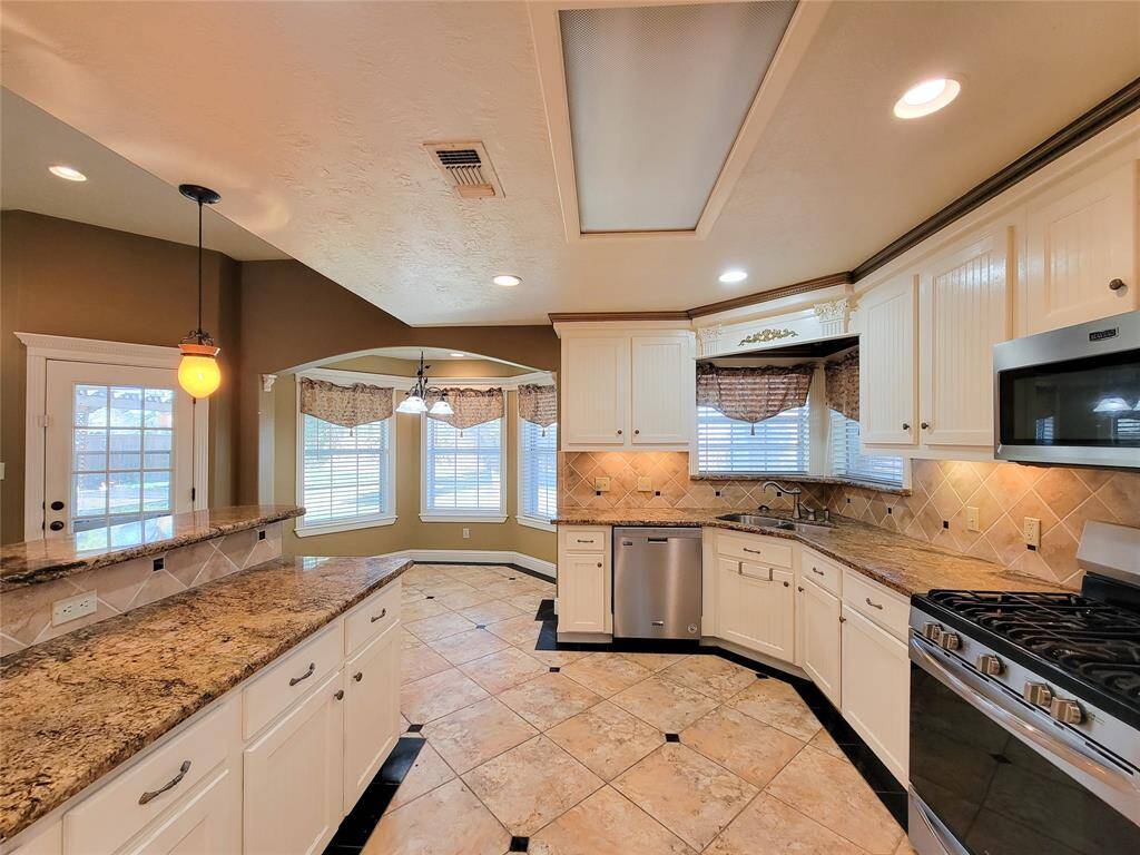 This kitchen features elegant granite countertops, white cabinetry, and stainless steel appliances. The space is well-lit with recessed lighting and a windowed dining nook, creating a warm and inviting atmosphere. The tile flooring and backsplash add a touch of sophistication, while the pendant light above the bar area enhances the room's charm.