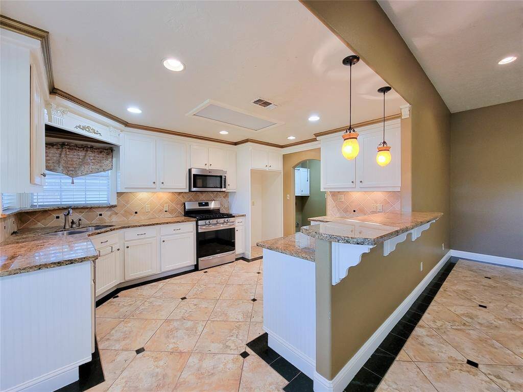 This is a spacious kitchen featuring white cabinetry, granite countertops, a tile backsplash, and modern appliances. It includes a breakfast bar with pendant lighting and tiled flooring throughout.