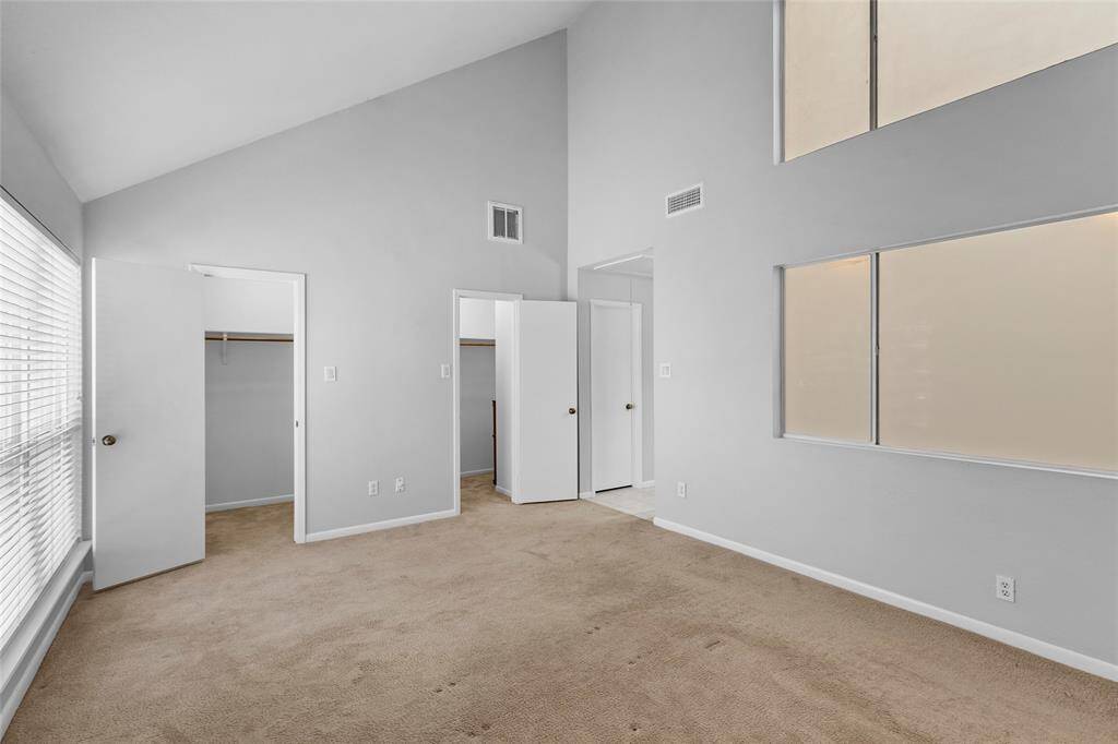 Here is your spacious primary bedroom without virtual staging, featuring neutral carpet and paint, high ceilings, large windows that fill the space with natural light, and a walk-in closet. Ideal for a bedroom or office.