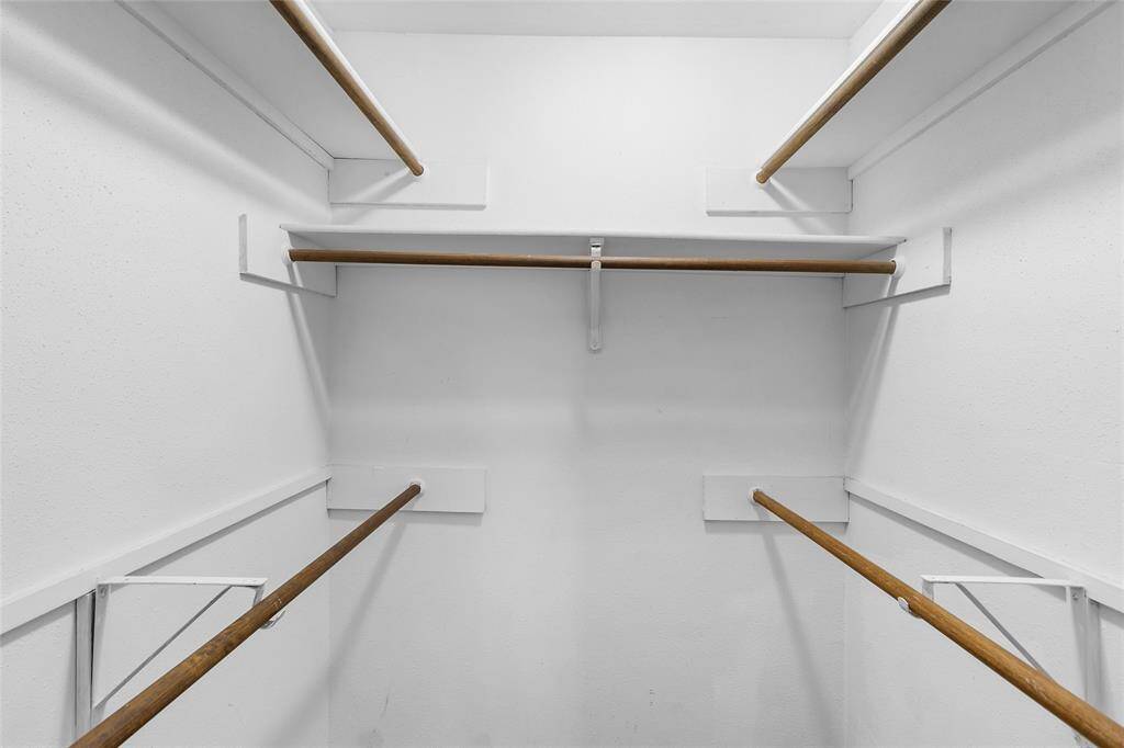 Here is your first primary walk-in closet, a spacious storage area featuring multiple wooden hanging rods and crisp white walls. This well-designed space offers plenty of room to keep your wardrobe organized and easily accessible.