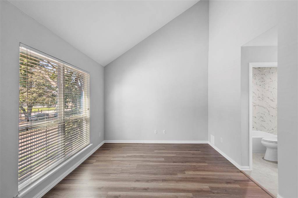 Here is another look at your spacious secondary bedroom, featuring high ceilings, neutral walls, and a large window that provides ample natural light. It has wood-like flooring and an adjacent bathroom with modern finishes. This is the photo without virtual staging, offering a true view of the room’s potential.