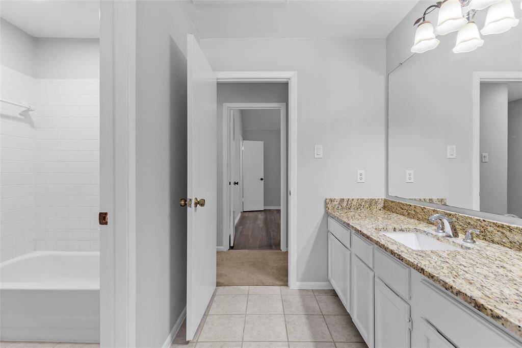 Your bathroom blends modern design with practical comfort. It features a sleek granite countertop with a single sink, a large mirror, and ample cabinet space for storage. Neutral-colored walls and tiled flooring keep the space feeling bright and inviting, while a glimpse of the bathtub/shower area adds a touch of spa-like relaxation.