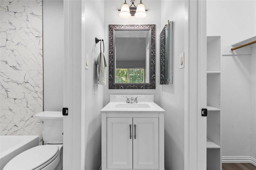 This is a compact, modern bathroom featuring a stylish vanity with an ornate mirror and overhead lighting. It includes a toilet and a shower with marble-patterned tiles. The space is bright and clean, with neutral colors and a small window for natural light.