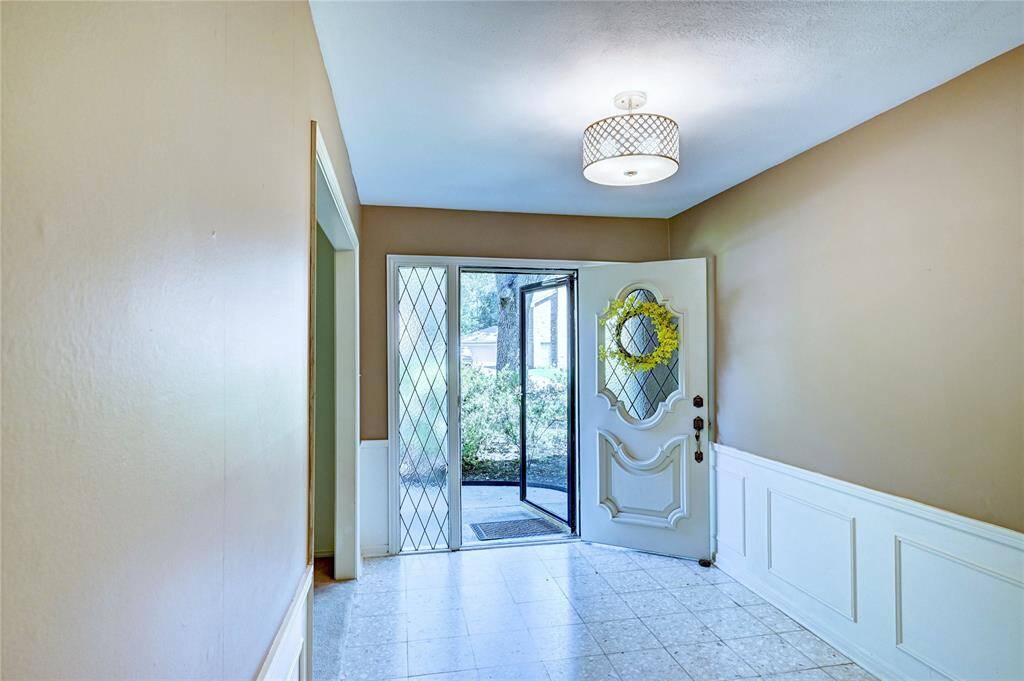 This inviting entryway features a classic white door with decorative glass panels, a welcoming wreath, and neutral walls. The space is brightened by a modern light fixture and offers a view of the lush greenery outside.