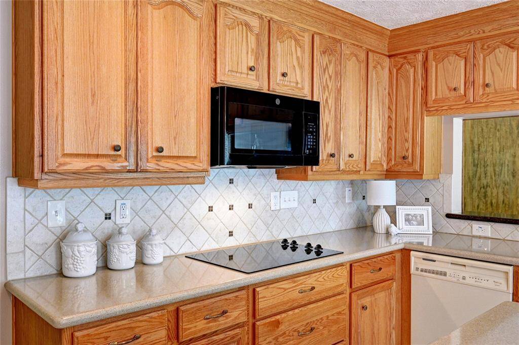 This kitchen features warm wooden cabinetry, a sleek black microwave, and an electric cooktop. The light-colored countertop and tiled backsplash add brightness, complemented by a built-in dishwasher and decorative accents for a cozy, functional space.