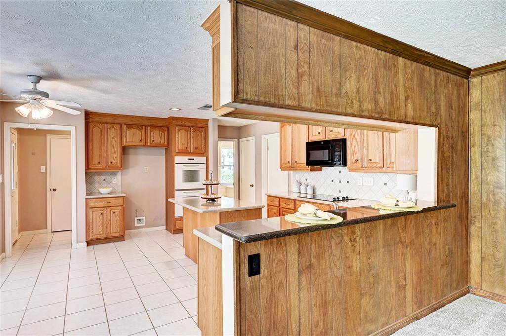 Spacious kitchen featuring warm wood cabinetry and a tiled backsplash. It includes modern appliances, ample counter space, and a convenient breakfast bar. The open layout connects seamlessly to the adjacent room, and the ceiling fan adds comfort.