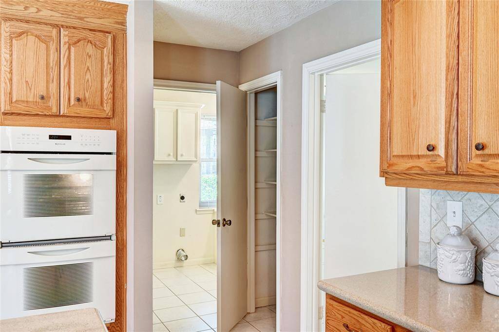 This kitchen features warm wooden cabinetry, a double oven, and tiled backsplash. There's a view into a separate utility room with ample shelving, offering convenient laundry access.