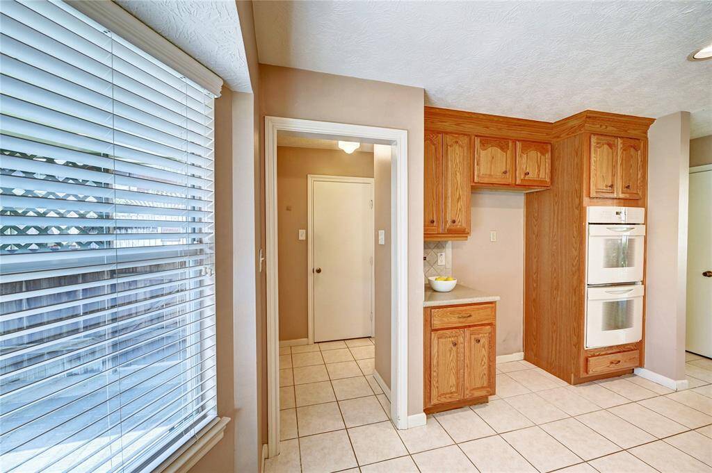 Bright kitchen with large windows, tiled floors, and wooden cabinetry. Features a built-in double oven and ample counter space, creating a warm and functional cooking area.