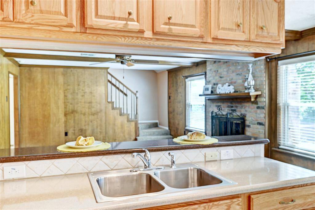 Kitchen with a view into the living area. The kitchen features wooden cabinets, a dual sink, and a countertop with a serving window. The living space includes a brick fireplace, wood paneling, and large windows for natural light. The staircase adds a cozy, homely touch.