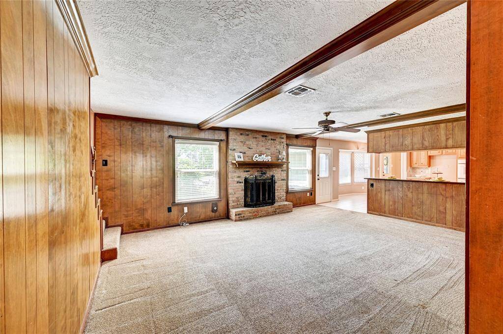 This spacious living room features wood paneling, a brick fireplace, and an open layout connecting to the kitchen. The room has a cozy feel with carpeted flooring and ample natural light from large windows.