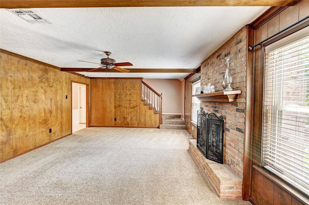 This cozy living room features warm wood paneling, a brick fireplace with a decorative mantel, a ceiling fan, and carpeted floors. Large windows bring in natural light, and a staircase leads to the upper level.