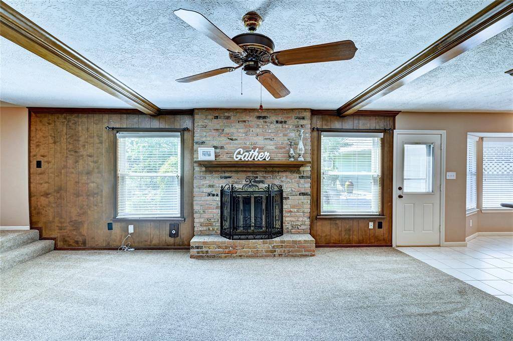 Spacious living room with cozy carpet and a central brick fireplace, featuring wood paneling and ceiling beams. Two large windows provide natural light, complemented by a ceiling fan for comfort.