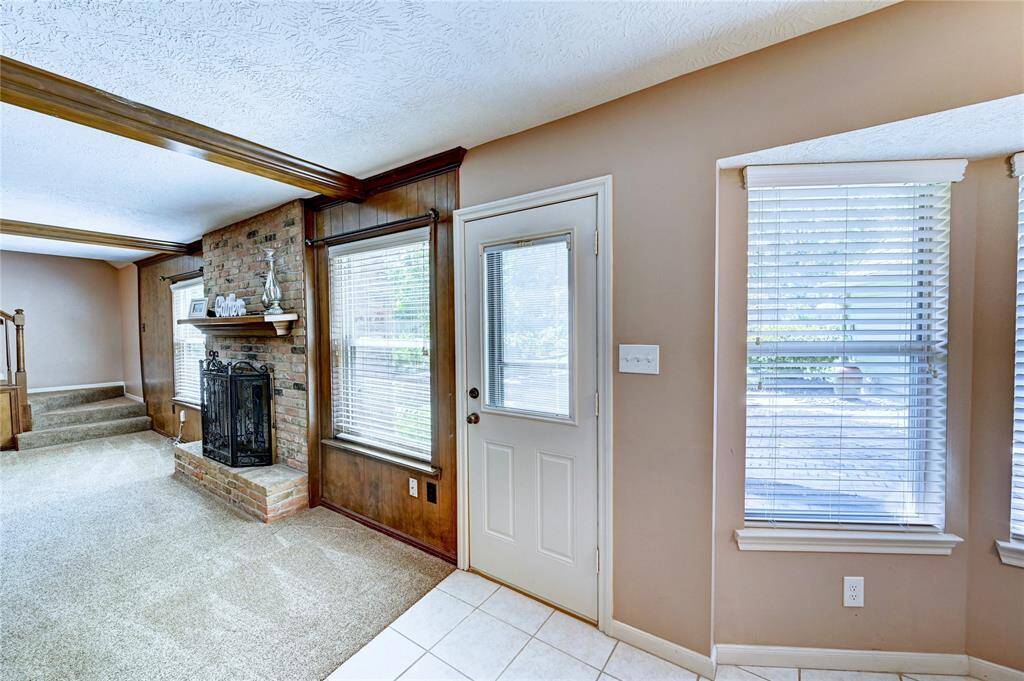 Cozy living area with a brick fireplace, carpeted flooring, and ample natural light from large windows. Features wood paneling and ceiling beams, with a tile entryway leading to an outdoor space.