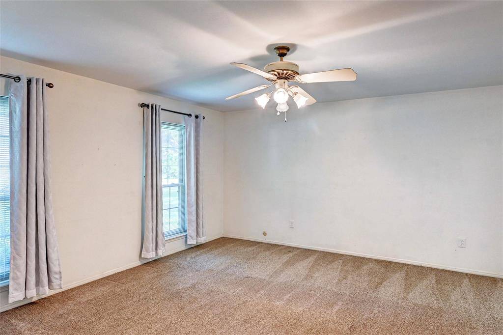 This room features neutral carpet and walls, two windows with curtains, and a ceiling fan with lights, offering a bright and airy space.