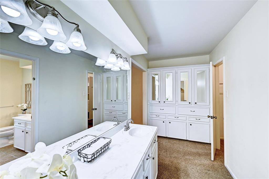 This photo showcases a bright, spacious bathroom with a sink, ample counter space, and abundant cabinetry for storage. The room is well-lit with stylish overhead lighting, and there's a glimpse of an adjoining room.