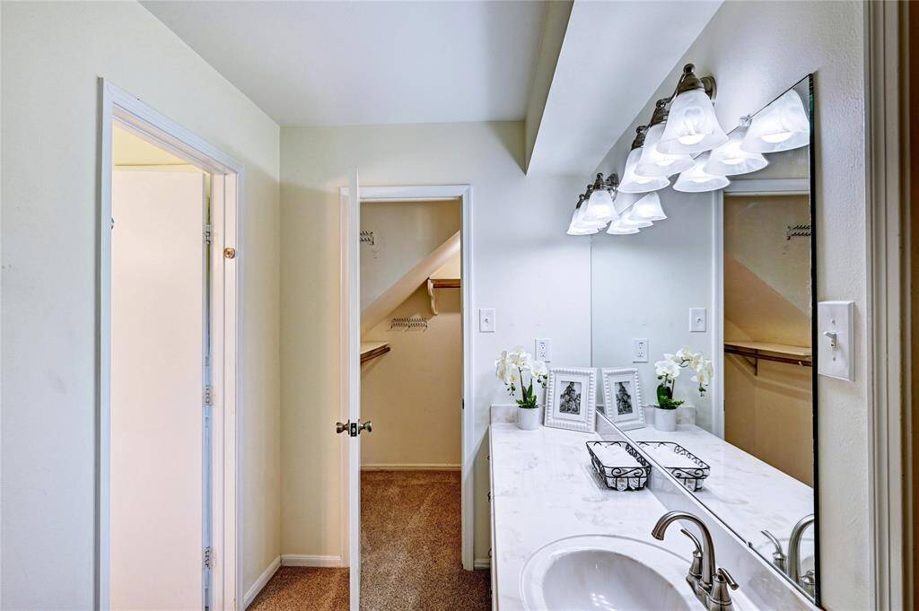 Bright bathroom with a white vanity, a large mirror, and elegant lighting. The space includes a sink with a modern faucet, decorative elements, and an adjacent walk-in closet with ample shelving.