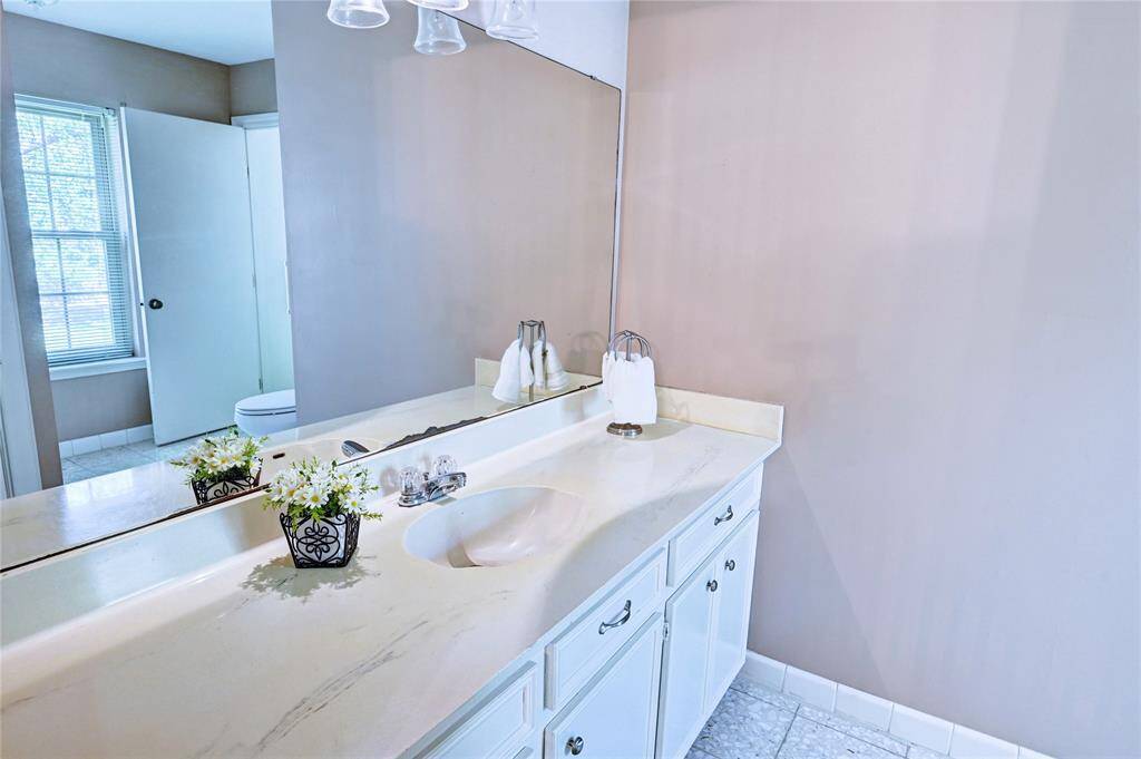 This bathroom features a sleek countertop with a built-in sink, a large mirror, and ample cabinet storage. Natural light enters through a window, enhancing the bright, welcoming atmosphere.