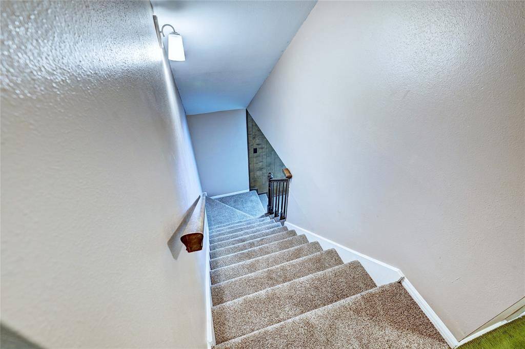 Carpeted staircase with a wooden handrail, neutral walls, and a modern wall light fixture, leading to a lower level.