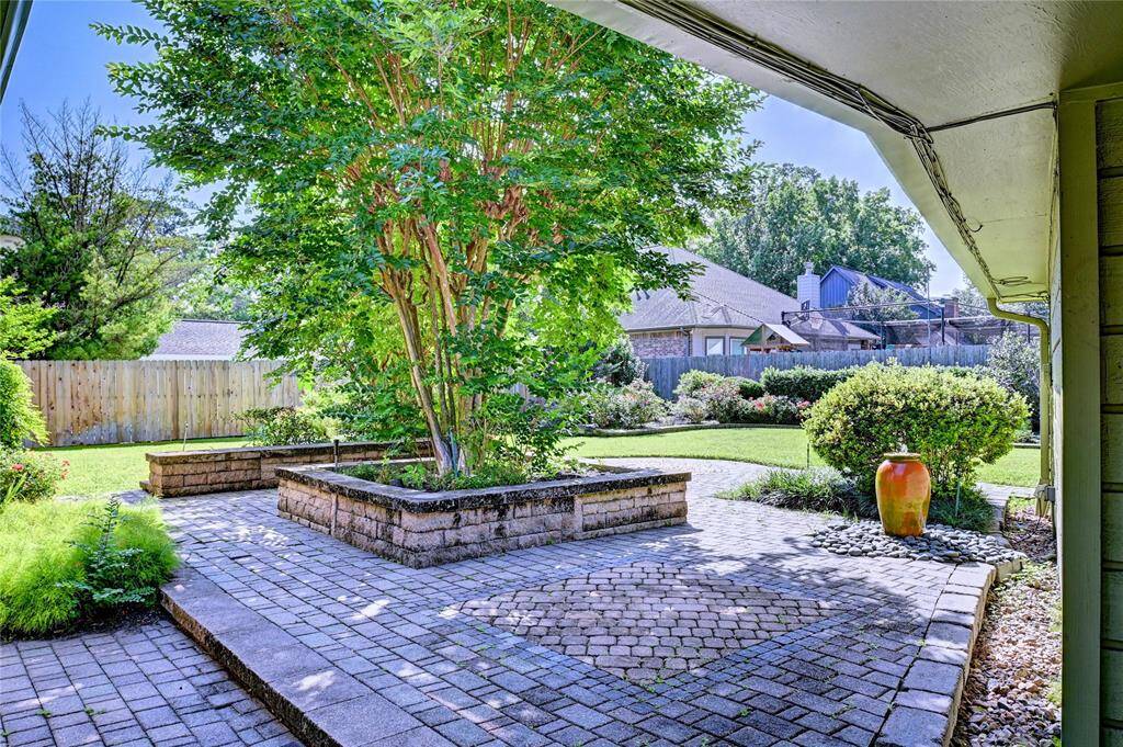 This charming backyard features a spacious, well-maintained patio with elegant pavers and a central raised planter bed with lush greenery. The surrounding garden is vibrant, offering a serene and private outdoor space ideal for relaxation or entertaining.