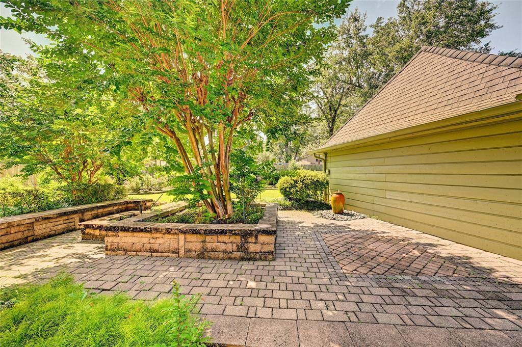 Peaceful backyard with a spacious brick patio, lush greenery, and mature trees. The area includes a well-maintained garden bed and offers a serene outdoor space perfect for relaxation or entertaining.