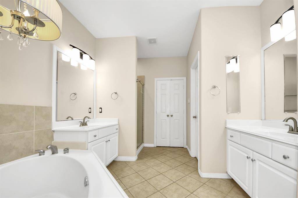 This bright bathroom features dual vanities with large mirrors, a spacious bathtub, neutral tiles, and a walk-in shower, offering a modern and functional space.